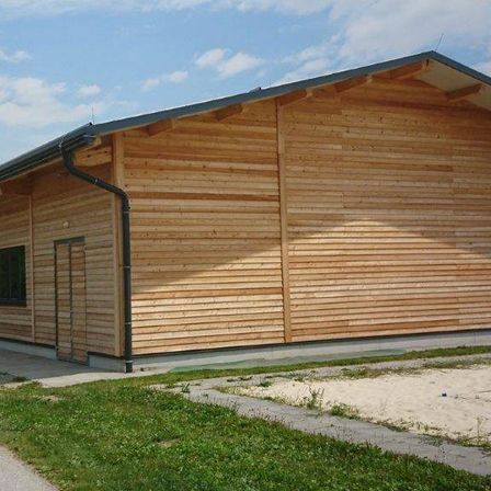 Ein hölzernes Gebäude mit einem grauen Dach und einem schwarzen Dachrinnenrohr. Das Äußere besteht aus Holzbrettern. Es gibt zwei Fenster auf der linken Seite. Vor dem Gebäude befindet sich eine Rasenfläche.
