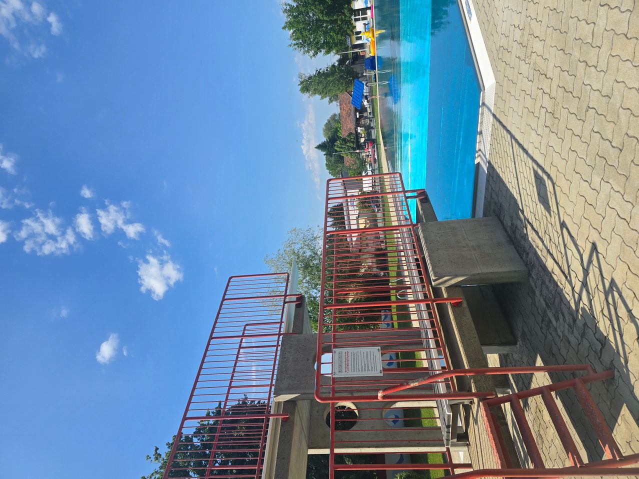 Ein großer Swimmingpool mit blauem Wasser und einem roten Metallgeländer, vor dem Hintergrund von Bäumen und einem klaren blauen Himmel.