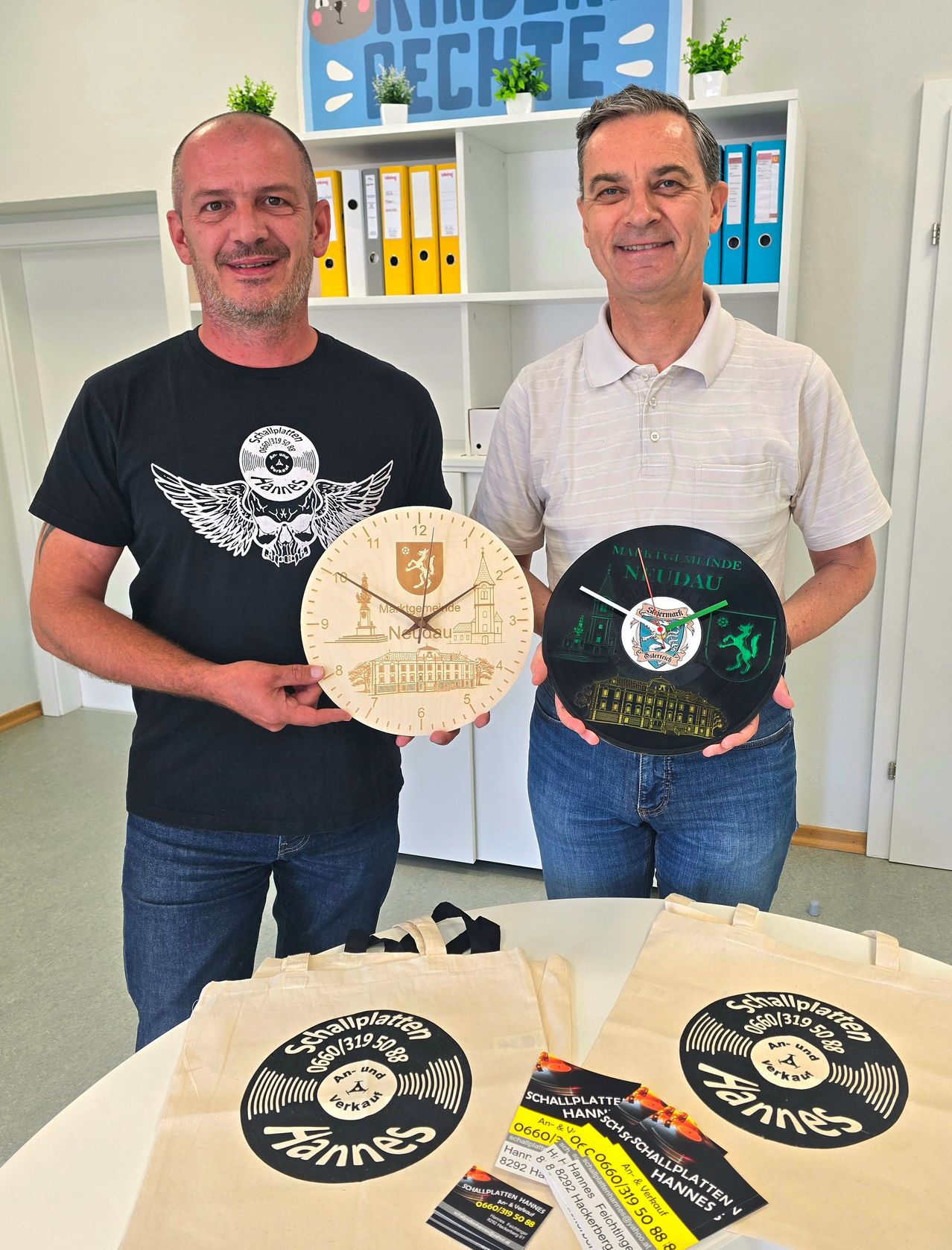 Zwei Männer halten runde Uhren. Ein Mann trägt ein schwarzes T-Shirt und der andere ein weißes Poloshirt. Sie stehen in einem Büro mit Regalen. Taschen mit Schallplatten-Text stehen auf dem Tisch.