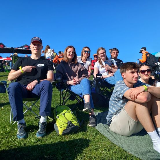 Eine Gruppe von Menschen sitzt auf Stühlen auf einem Grasfeld unter einem klaren blauen Himmel. Einige tragen eine Brille und Sneakers. Ein Mann vorne trägt einen schwarzen Hut und eine Armband.