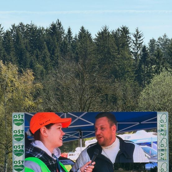 Eine Frau mit einer roten Kappe und ein Mann in einem weißen Shirt sprechen in ein Mikrofon unter einem blauen Zelt, mit Bäumen und einem großen Bildschirm im Hintergrund.