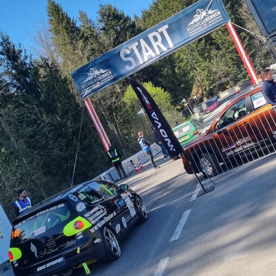 Ein Auto mit vielen Aufklebern fährt unter einem Banner mit der Aufschrift 'Start'. Mehrere Personen stehen herum, und Autos sind hinter Barrieren geparkt.
