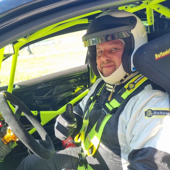 Ein Mann in einem Rennanzug sitzt in einem gelben und grünen Auto. Er trägt einen Helm, Handschuhe und eine Nackenstütze. Er lächelt und gibt ein Daumen hoch.
