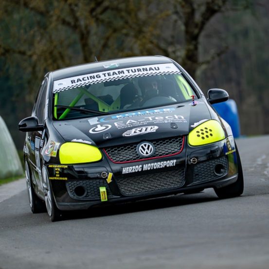 Ein schwarz-gelber Volkswagen Golf GTI fährt auf einer Rennstrecke mit Racing Team Turnau und Herzog Motorsport auf der Front.