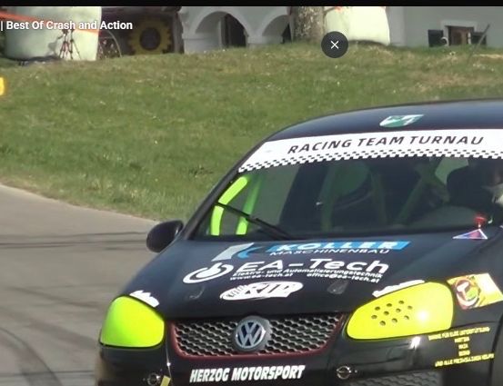 Ein schwarzer Rennwagen mit grünen Akzenten fährt auf einer Rennstrecke mit Gras am Rand und einem weißen Gebäude im Hintergrund.