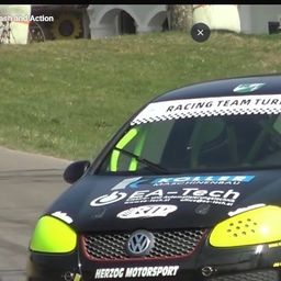 Ein schwarzer Rennwagen mit grünen Akzenten fährt auf einer Rennstrecke mit Gras am Rand und einem weißen Gebäude im Hintergrund.