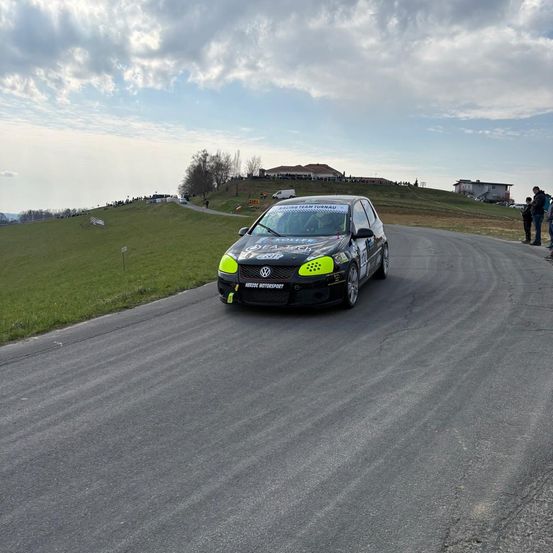 Ein Rennwagen fährt auf einer Straße mit einem grün bewachsenen Hügel und einigen Gebäuden in der Ferne. Das Auto ist schwarz mit grünen Akzenten und hat die Nummer 1 auf der Vorderseite.