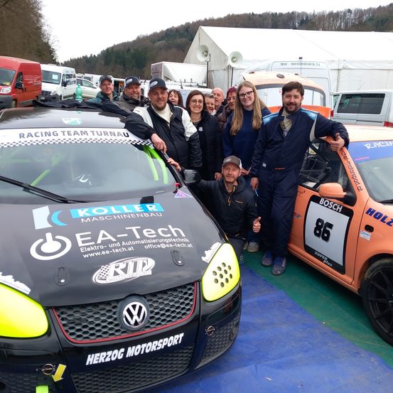 Eine Gruppe von Menschen steht hinter zwei Rennwagen. Das schwarze Auto hat 'Racing Team Turnau' auf der Motorhaube. Das orangefarbene Auto hat '86' an der Seite. Sie tragen alle Rennanzüge.