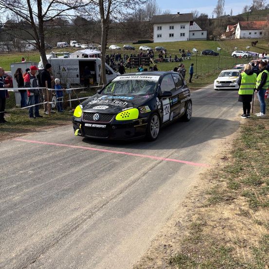Ein schwarzer und gelber Wagen fährt auf einer Straße mit Zuschauern. Bäume, Häuser und Autos säumen die Seite. Ein weißer Van ist in der Nähe geparkt. Leute schauen von hinter einer Barriere zu.