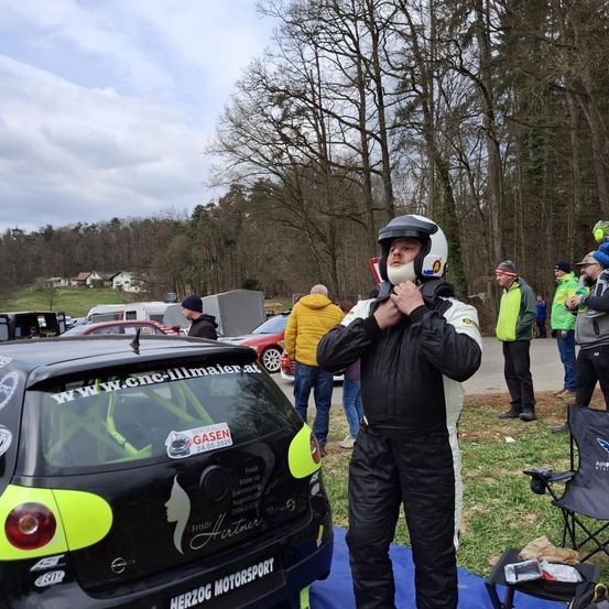 Ein Mann in Helm und Rennanzug steht neben einem schwarzen Auto mit 'Herzog Motorsport' auf der Rückseite. Mehrere Personen sind im Hintergrund, einige stehen in der Nähe von Autos.