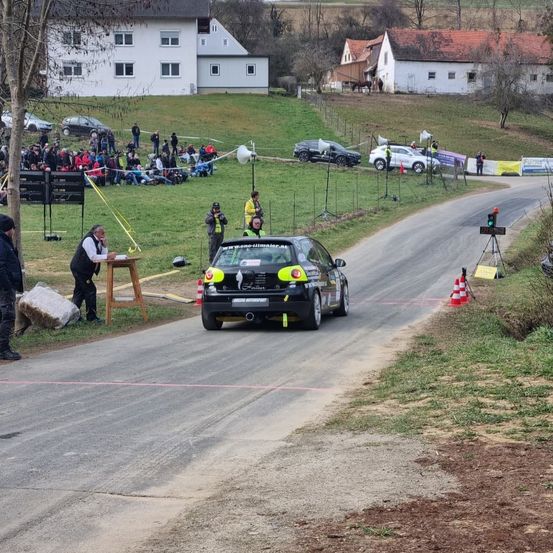 Ein Auto fährt auf einer kurvenreichen Straße, mit Zuschauern, die sich an der Seite versammelt haben. Ein Mann steht in der Nähe eines Tisches und einer Barriere. Es gibt Verkehrskegel und einen Pfosten mit einer Kamera. Bäume und Häuser sind im Hintergrund zu sehen.