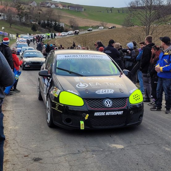 Eine Gruppe von Zuschauern beobachtet ein Rennauto mit der Aufschrift 'Racing Team Turnau' auf der Motorhaube. Das Auto steht auf einer Straße mit anderen Autos, und die Menschen tragen Winterkleidung.