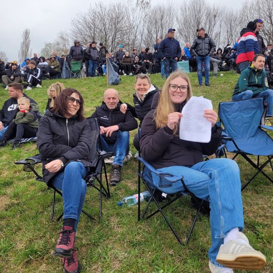 Eine Gruppe von Menschen sitzt auf Klappstühlen auf einem grasbewachsenen Hügel. Eine Frau hält ein Papier, und ein Mann neben ihr lächelt. Im Hintergrund stehen und sitzen viele Menschen auf dem Gras.