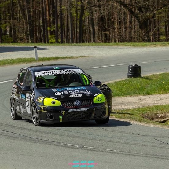 Ein schwarz-grüner Rennwagen fährt auf einer Kurvenbahn. Der Wagen hat die Aufschrift 'Racing Team Turnau' auf der Motorhaube. Der Wagen ist von Gras und einem Mülleimer umgeben.
