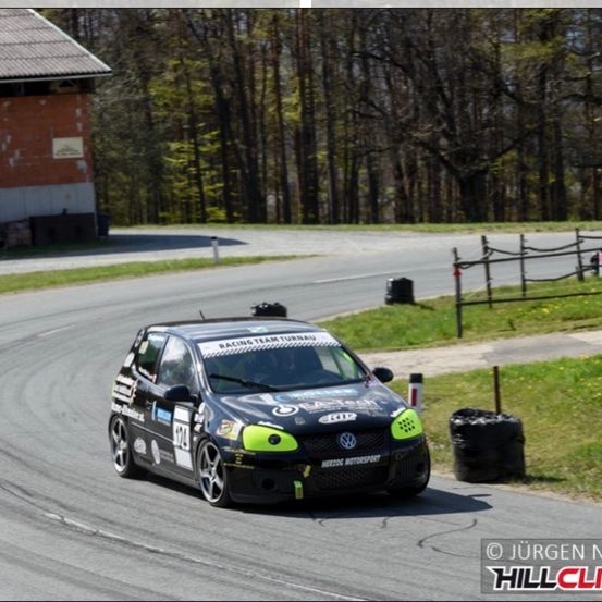 Ein schwarzer Volkswagen Polo GTI Rennwagen fährt eine Kurve auf einer Rennstrecke. Bäume und Gras säumen die Ränder, im Hintergrund ist ein Gebäude und ein Zaun zu sehen.