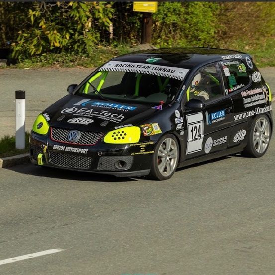 Ein schwarz-gelber Wagen mit Racing Team Turnau-Aufklebern fährt auf einer Straße, mit Bäumen und einem Schild im Hintergrund.