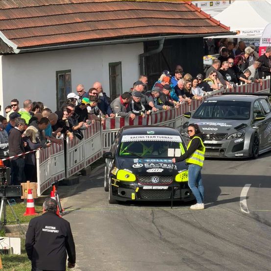 Ein Rennwagen steht auf der Rennstrecke, eine Frau in einer gelben Weste steht daneben. Zuschauer beobachten von einer Absperrung aus. Ein Gebäude mit einem braunen Dach ist im Hintergrund.