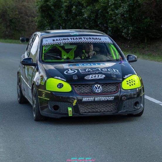 Ein schwarzer Volkswagen Golf mit grünen Akzenten, gesponsert von Herzog Motorsport, fährt auf einer Landstraße mit Bäumen an den Seiten.