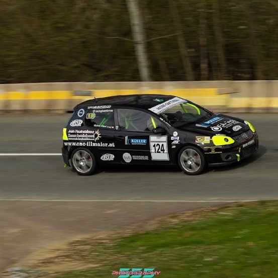 Ein schwarzer Rennwagen mit Sponsorenlogo fährt auf einer Rennstrecke mit verschwommenem Hintergrund. Das Auto hat die Nummer 124 an der Seite und gelbe Akzente.