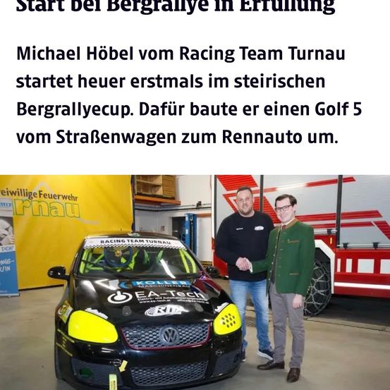 Zwei Männer stehen neben einem Rennwagen in einer Garage, einer trägt einen grünen Anzug, sie schütteln Hände. Das Auto hat ein gelbes und schwarzes Design mit den Worten EA Tech. Hinter ihnen steht ein Feuerwehrwagen.