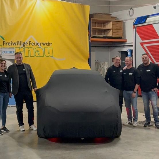 Fünf Personen stehen um ein in Schwarz verhülltes Auto. Der Mann links trägt eine Jacke, die anderen schwarze Hemden. Ein gelber Banner steht hinter ihnen.