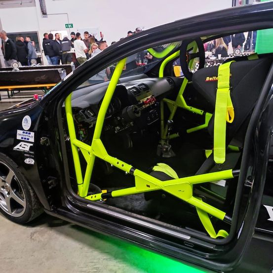 Ein schwarzer Wagen mit neongrünen Rennzubehörteilen ist zu sehen, mit geöffneter Tür. Das Interieur verfügt über einen Rennsitz und ein Lenkrad mit Sicherheitsgurten. Im Hintergrund beobachten Menschen.