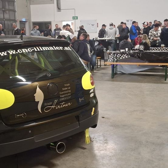 Die Rückseite eines schwarzen Autos mit gelben Akzenten steht in einer Garage. Es gibt versammelte Menschen und Tische mit Gegenständen sind aufgebaut. Das Auto hat einen Aufkleber mit Logo und Text.