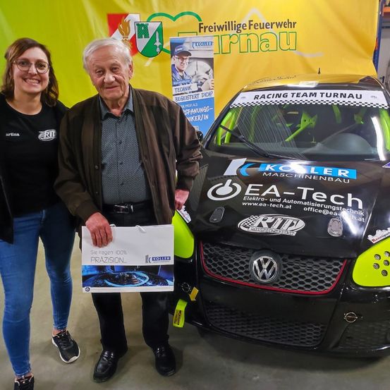Ein älterer Mann steht neben einer Frau, beide lächeln, vor einem grünen und schwarzen Rennwagen. Der Mann hält einen weißen Geschenkbeutel. Der Wagen trägt den Text 'Racing Team Turnau'. Hinter ihnen zeigt ein Banner das Logo der Freiwilligen Feuerwehr Turnau.