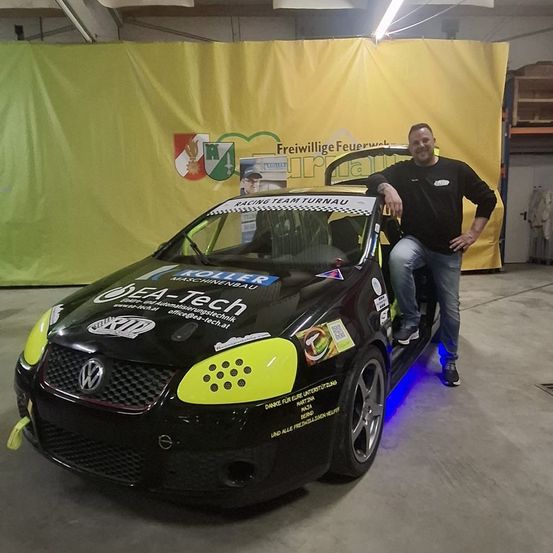 Ein Mann lehnt an einem schwarz-gelben Auto in einer Garage. Das Auto hat Logos und Text darauf. Ein gelber Vorhang mit einem Logo ist hinter dem Auto.