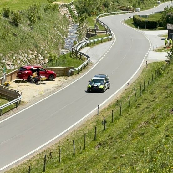 Ein rotes Auto parkt am Rande einer kurvenreichen Straße, in der Nähe zweier Personen. Ein grünes Auto fährt die Straße entlang.