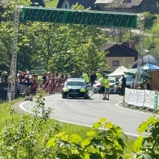 Eine Herzog Motorsport Veranstaltung mit einem Auto auf der Straße, umgeben von Menschen und Zelten. Bäume und Häuser sind im Hintergrund.