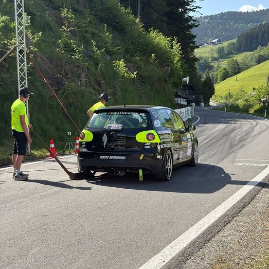 Ein Rennwagen fährt auf einer Straße, wobei zwei Personen in neongrünen Uniformen die Straße mit einem Besen reinigen.