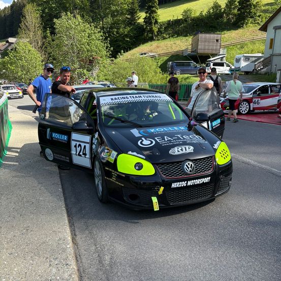Ein schwarz-grüner Rennwagen mit der Nummer 124 steht bei einem Rennevent. Zwei Männer stehen daneben. Bäume und Häuser sind im Hintergrund.