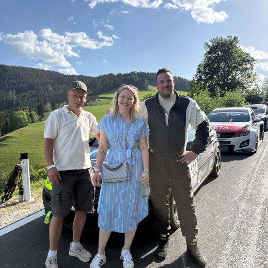 Drei Personen stehen vor einem Auto auf einer Straße mit Bergen im Hintergrund. Ein Mann links trägt eine Kappe und Turnschuhe. Eine Frau in einem Kleid hält eine Handtasche. Der Mann rechts trägt eine Jacke und hält eine Flasche.