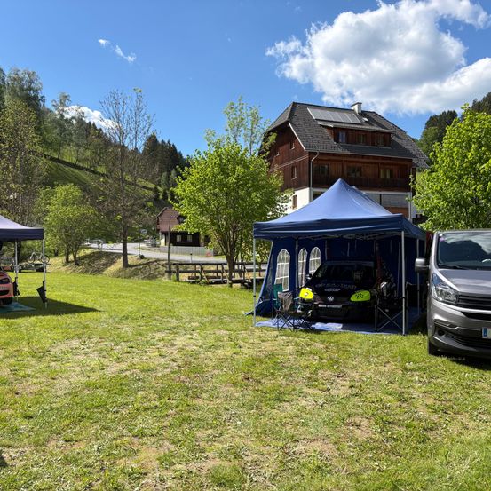 Ein Camping-Setup mit zwei Zelten und einem Auto unter einem blauen Zelt. Ein Haus und Bäume sind im Hintergrund.