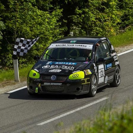 Ein schwarzer und grüner Wagen mit Racing Team Turnau Schriftzug auf der Motorhaube fährt auf einer Straße, im Hintergrund weht eine gepunktete Flagge.