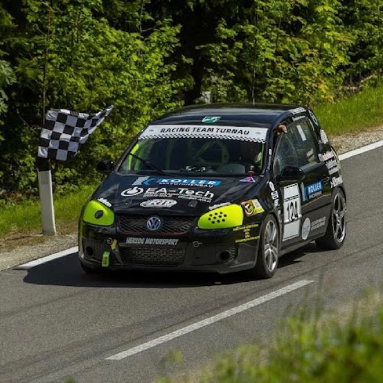 Ein schwarzer und grüner Wagen mit Racing Team Turnau Schriftzug auf der Motorhaube fährt auf einer Straße, im Hintergrund weht eine gepunktete Flagge.