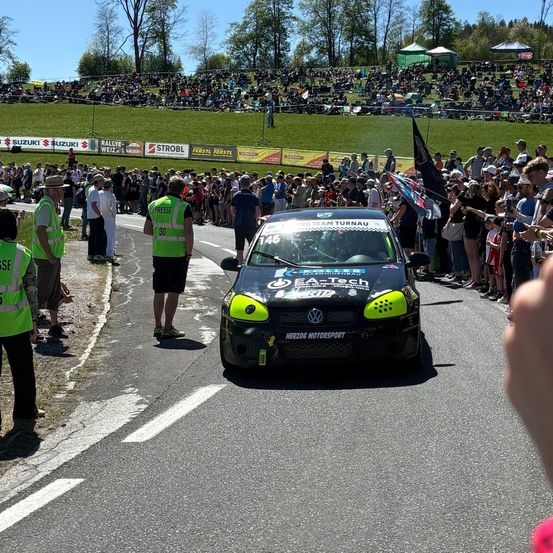 Ein Rallyeauto fährt auf der Straße, umgeben von einer großen Menschenmenge. Viele Menschen stehen an der Seite, einige tragen grüne Westen. Banner säumen die Straße, und eine große Anhöhe ist im Hintergrund zu sehen.