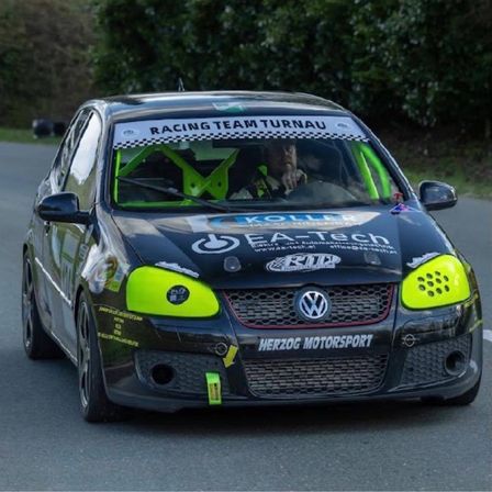 Ein schwarz-gelber Rennwagen mit Racing Team Turnau auf der Windschutzscheibe fährt auf einer Straße. Das Auto hat Herzog Motorsport auf dem Stoßfänger stehen.