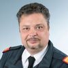 Ein Mann in Uniform lächelt, trägt eine Krawatte und einen Abzeichen mit roten, gelben und schwarzen Farben. Er hat graues Haar und einen Bart.