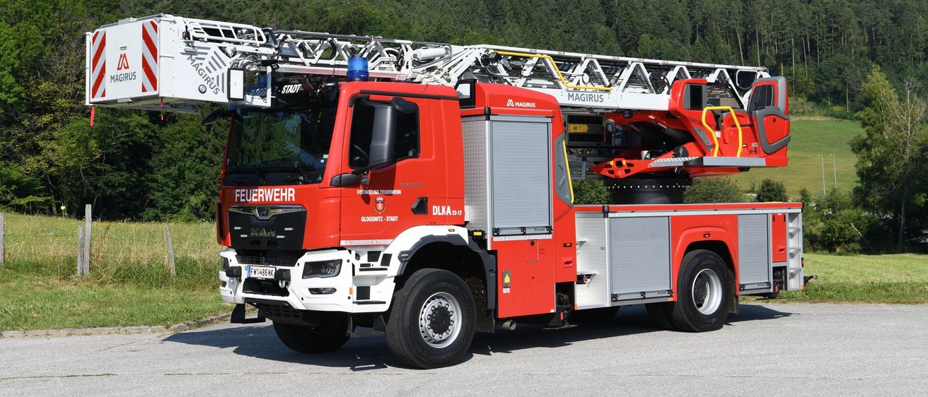 Ein roter Feuerwehrwagen mit einer Leiter steht auf der Straße, mit einem Grasfeld und einem Berg im Hintergrund.