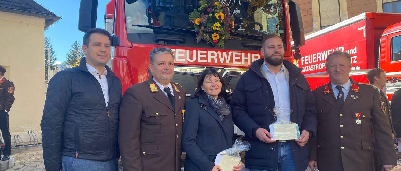 Fünf Personen posieren für ein Foto vor einem roten Feuerwehrauto mit Blumen. Sie halten ein Geschenk und tragen Winterkleidung. Das Gebäude dahinter hat rote Wände und Fenster.