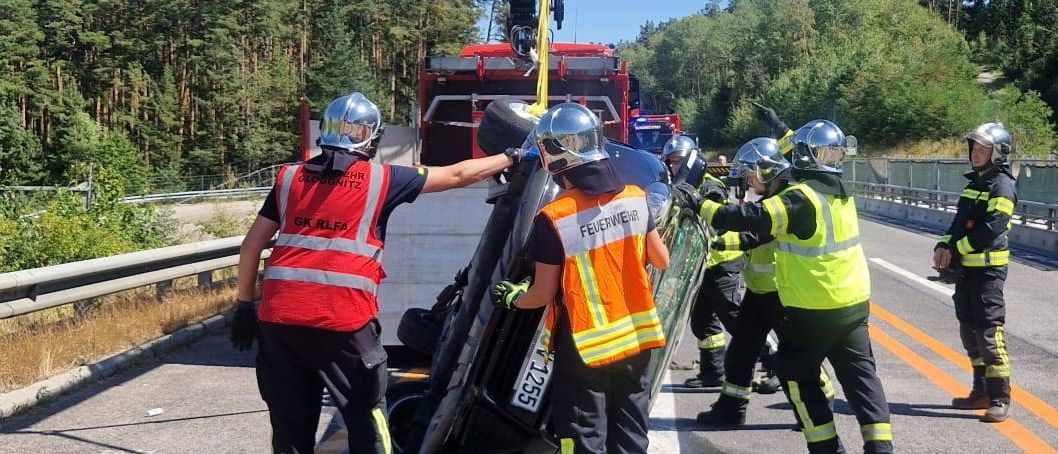 Einsatzkräfte in Warnwesten und Helmen heben ein beschädigtes Fahrzeug mit einem Kran von der Straße. Ein Feuerwehrwagen ist in der Nähe geparkt.