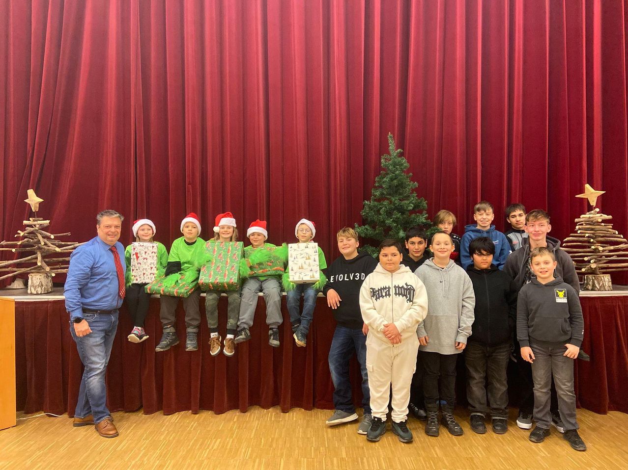 Eine Gruppe von Kindern, die grüne Weihnachtsmützen tragen und Geschenke halten, posieren mit einem Mann in einem blauen Hemd und Krawatte. Sie stehen hinter einem Weihnachtsbaum auf einer Bühne mit einem roten Vorhang.