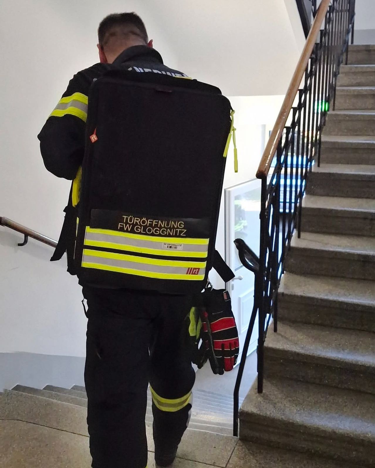 A firefighter is carrying a backpack with the words 'TUROFFNUNG FW GLOGGNITZ' on it. He is wearing a yellow and black safety jacket. He is standing on the stairs.