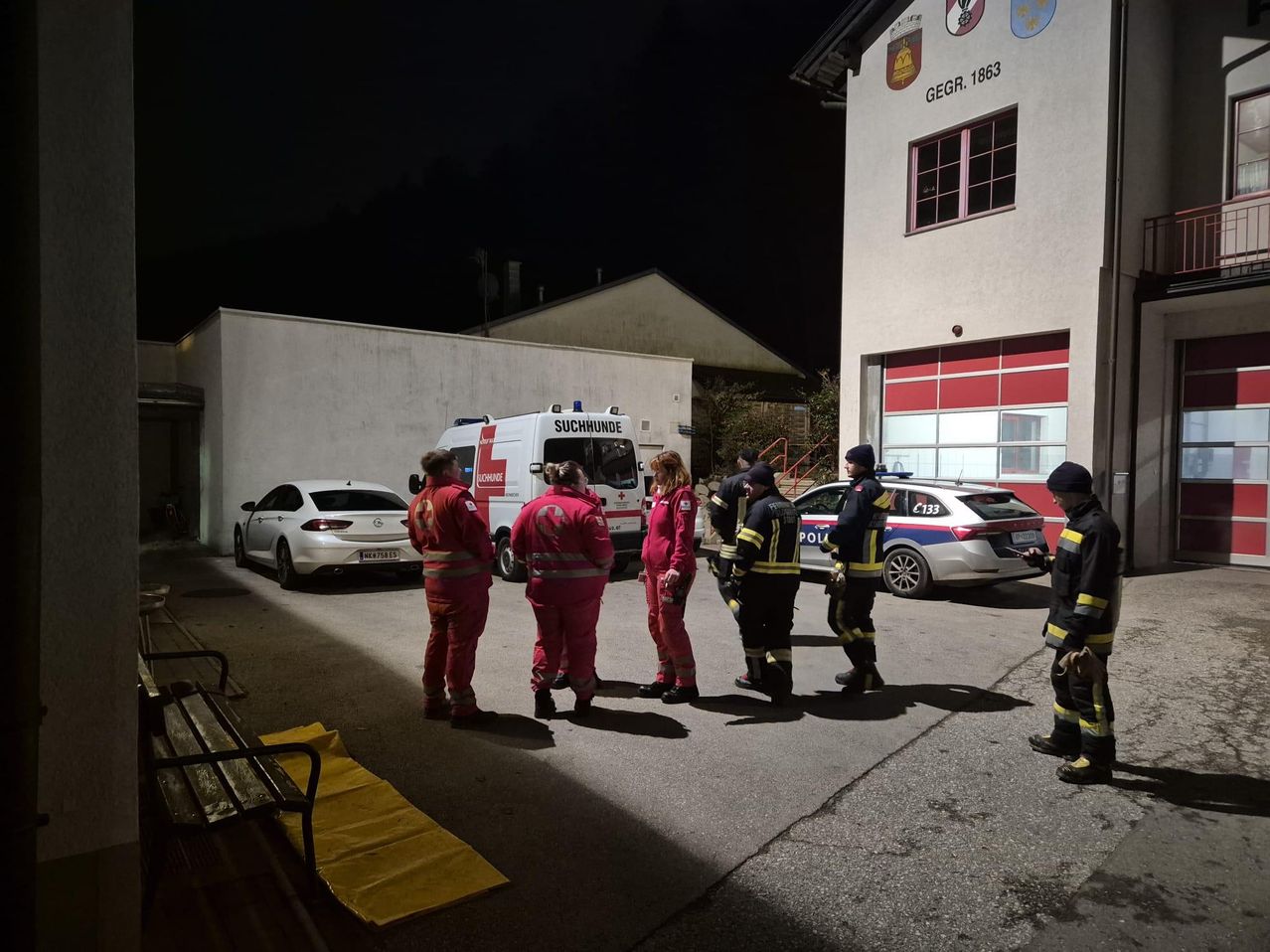 In der Nacht stehen mehrere Menschen in roten Uniformen vor einem Gebäude. Ein weißer Van und ein Polizeiauto sind vor dem Gebäude geparkt. Das Gebäude hat ein Logo an der Wand.