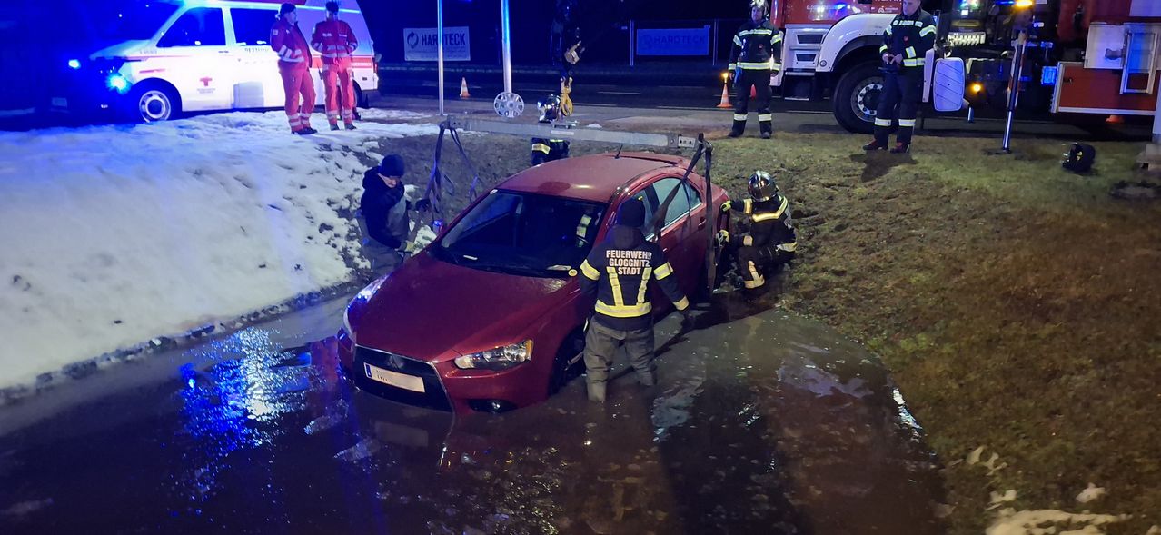 Feuerwehrleute retten ein in Wasser eingetauchtes rotes Auto. Zwei stehen beim Auto, einer klettert hinein und andere sind in der Nähe.