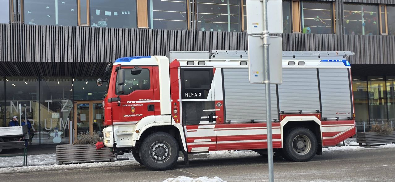 Ein rot-weißer Feuerwehrwagen mit der Aufschrift HLFA 3 steht an der Straßenseite. Der Wagen hat eine Leiter obenauf und steht vor einem Gebäude mit Glasfenstern.