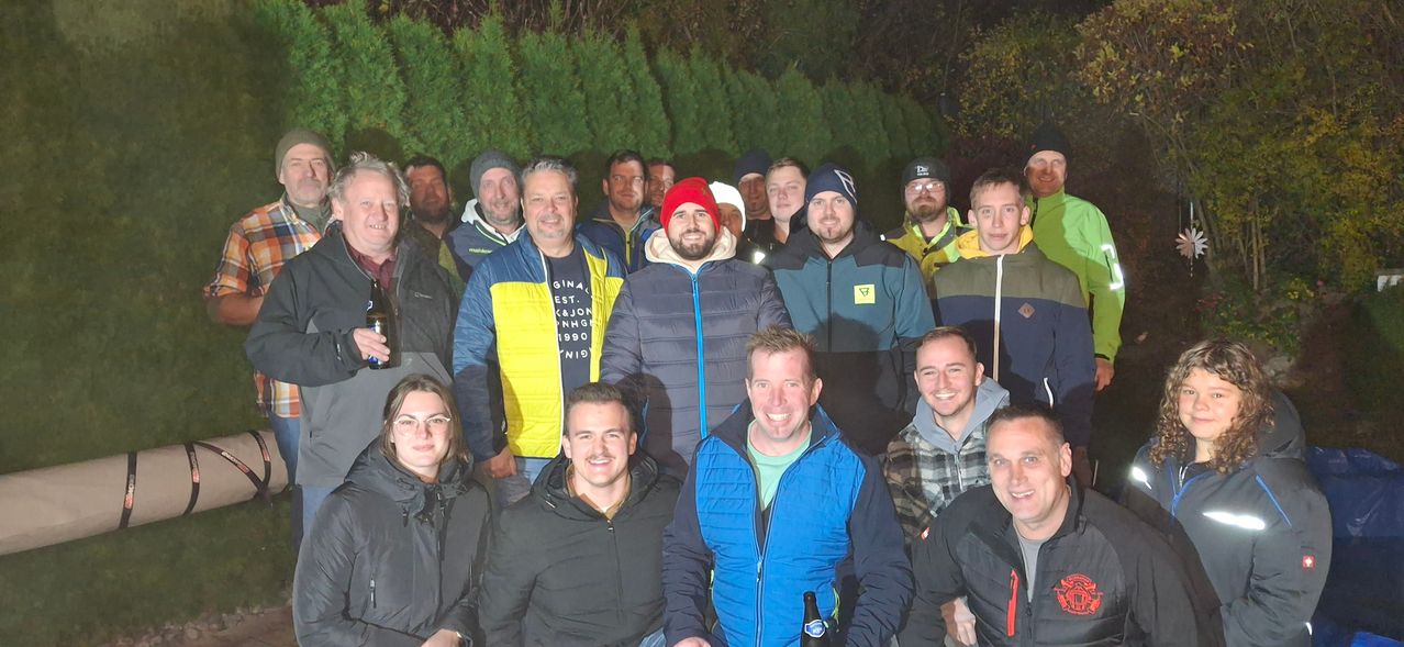 A group of people dressed in winter clothing are posing for a photo. They are smiling and appear to be enjoying themselves. The background is blurry with trees and foliage.