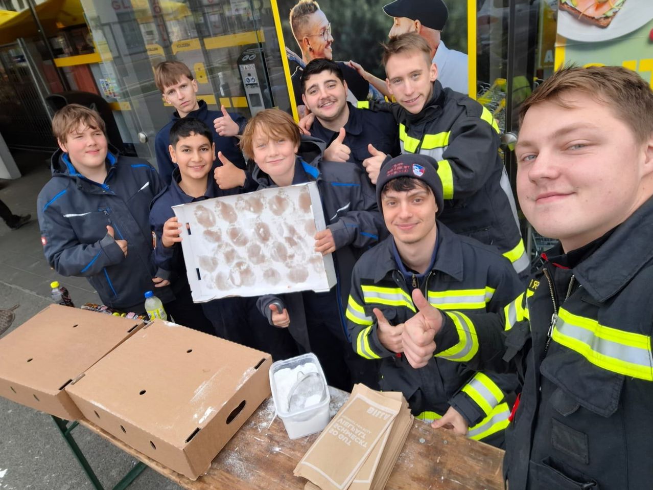 A group of firefighters and young boys are posing for a photo outside, with two cardboard boxes and a bag labeled 'BIFF' on a table.